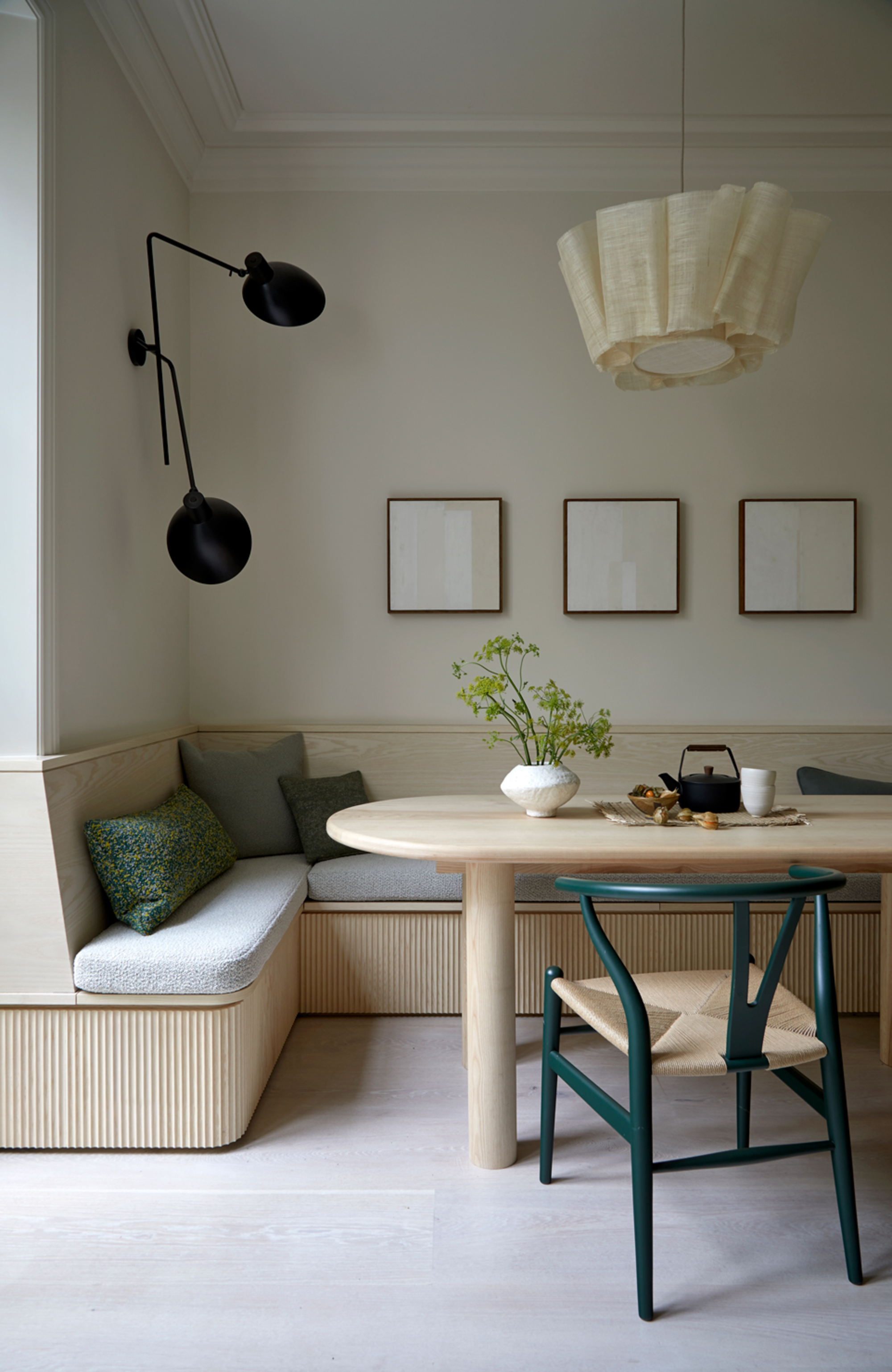 modern dining room with corner banquette seating with cushions, wall sconce, three artworks hanging on the wall, pendant hanging over the timber curved dining table with green wishbone chair