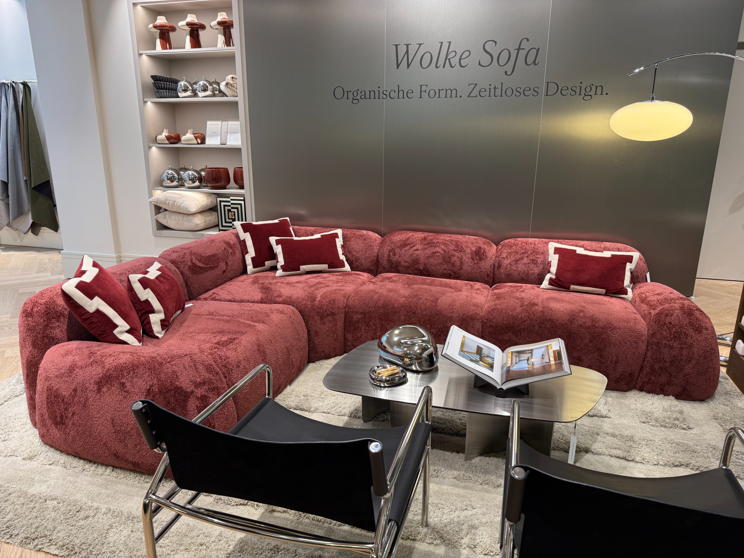 A large sectional wine red sofa with red scatter cushions with a coffee table and two sling back black chairs with steel frames in front of a sanded steel wall