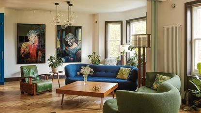 modern living room with blue sofa and green sofa and armchair with timber floors, large artworks, square timber coffee table, radiator on the wall, floor lamp, and pendant lights