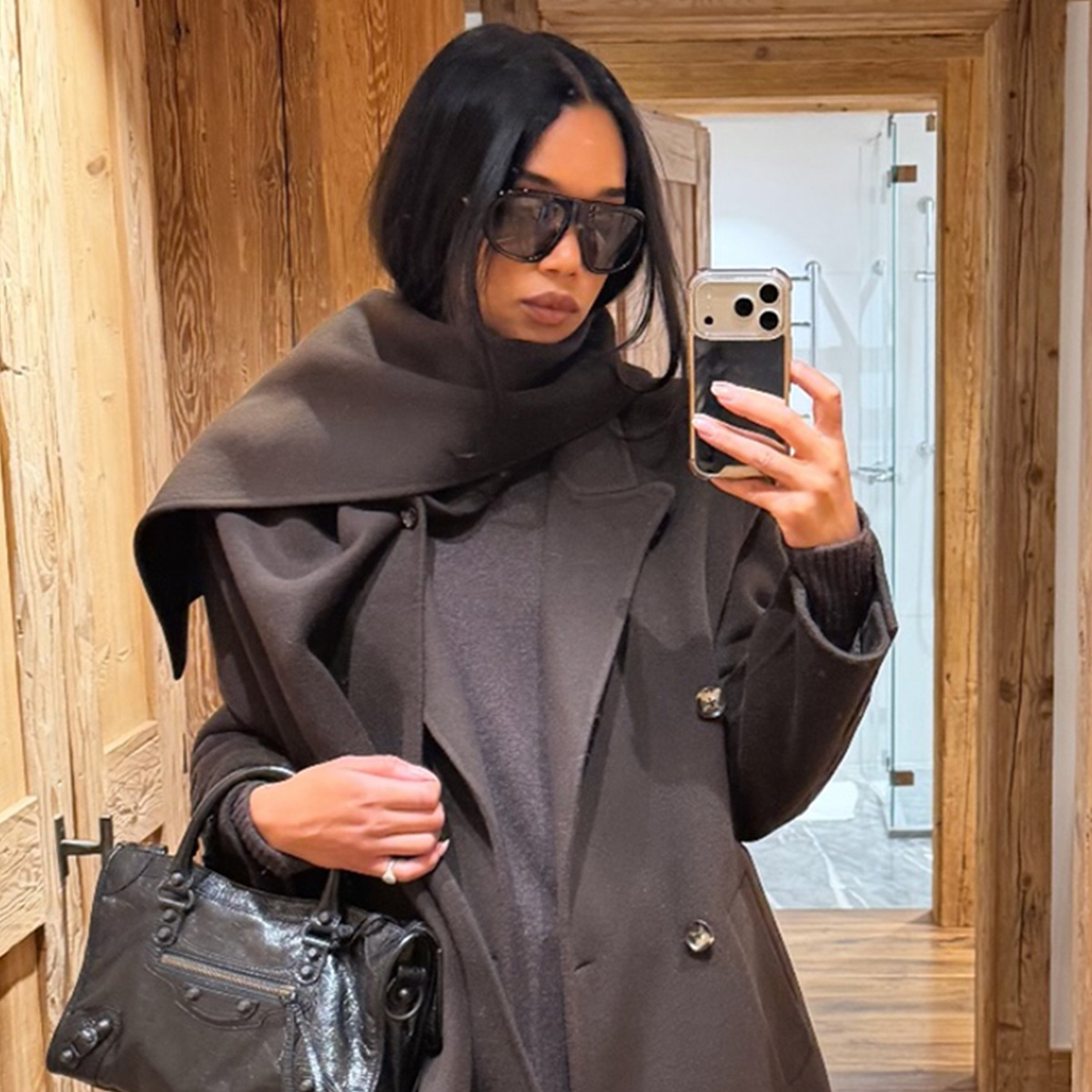 Laura Harrier wearing a brown coat, beige corduroy pants, and brown boots with a black Balenciaga City bag.