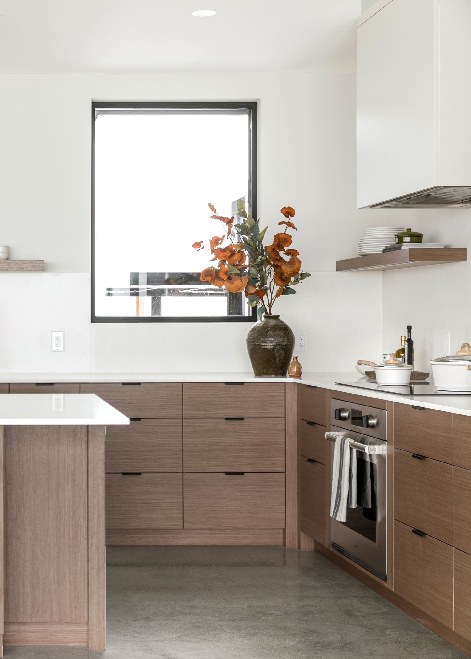 A wooden cabinets in a bright kitchen with a large window. A large vase sits by the window with flowers inside of it.