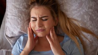 A woman lying in bed holding her jaw in pain after grinding her teeth in her sleep