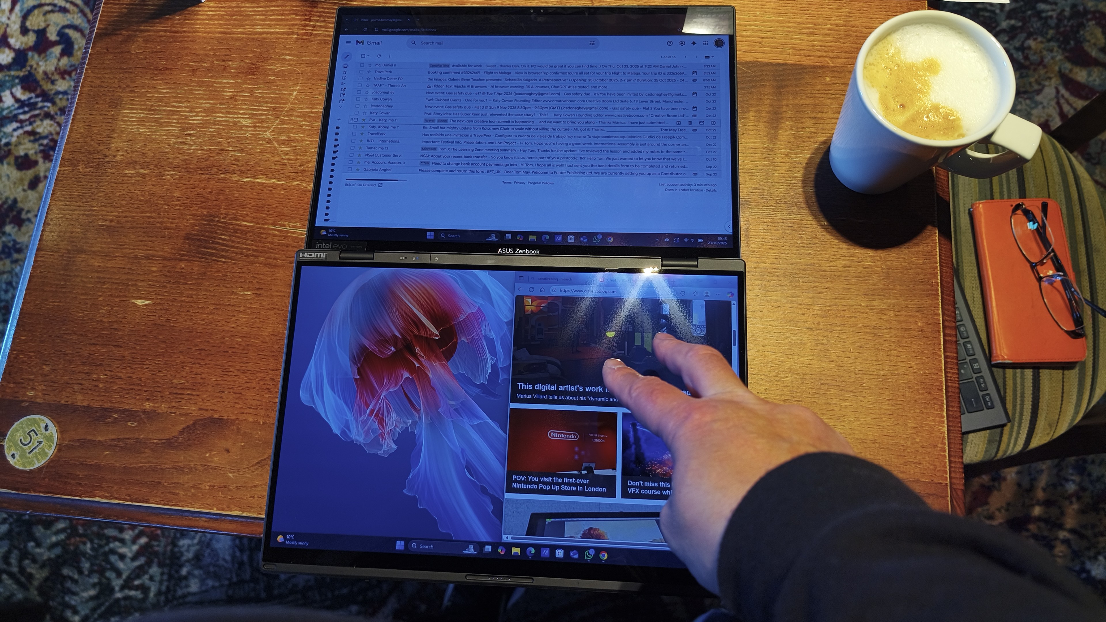 An overhead shot shows a dual-screen laptop laid flat on a wooden table, with a person's hand touching the lower screen, next to a coffee cup and a pair of glasses on a notebook.