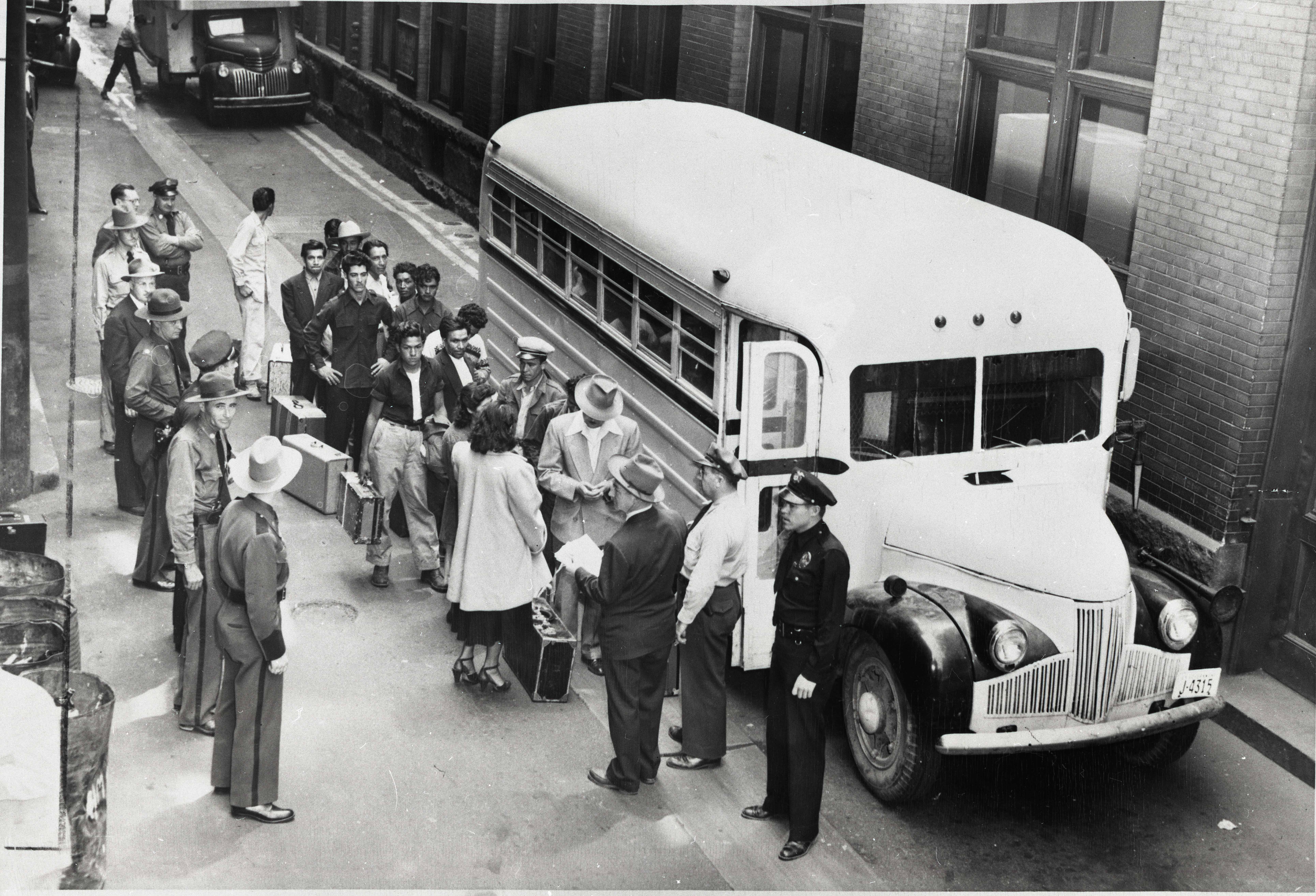 A line of men with suitcases wait beside a bus on a city street as police officers and officials stand nearby.