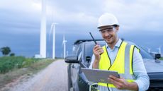 A field service technician wearing a high-vis vest looking at a tablet and speaking on their radio while laughing. Behind them, a car and a road flanked by wind turbines can be seen.