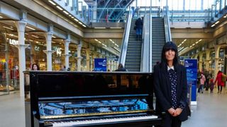 Claudia Winkleman stands beside a piano at a train station