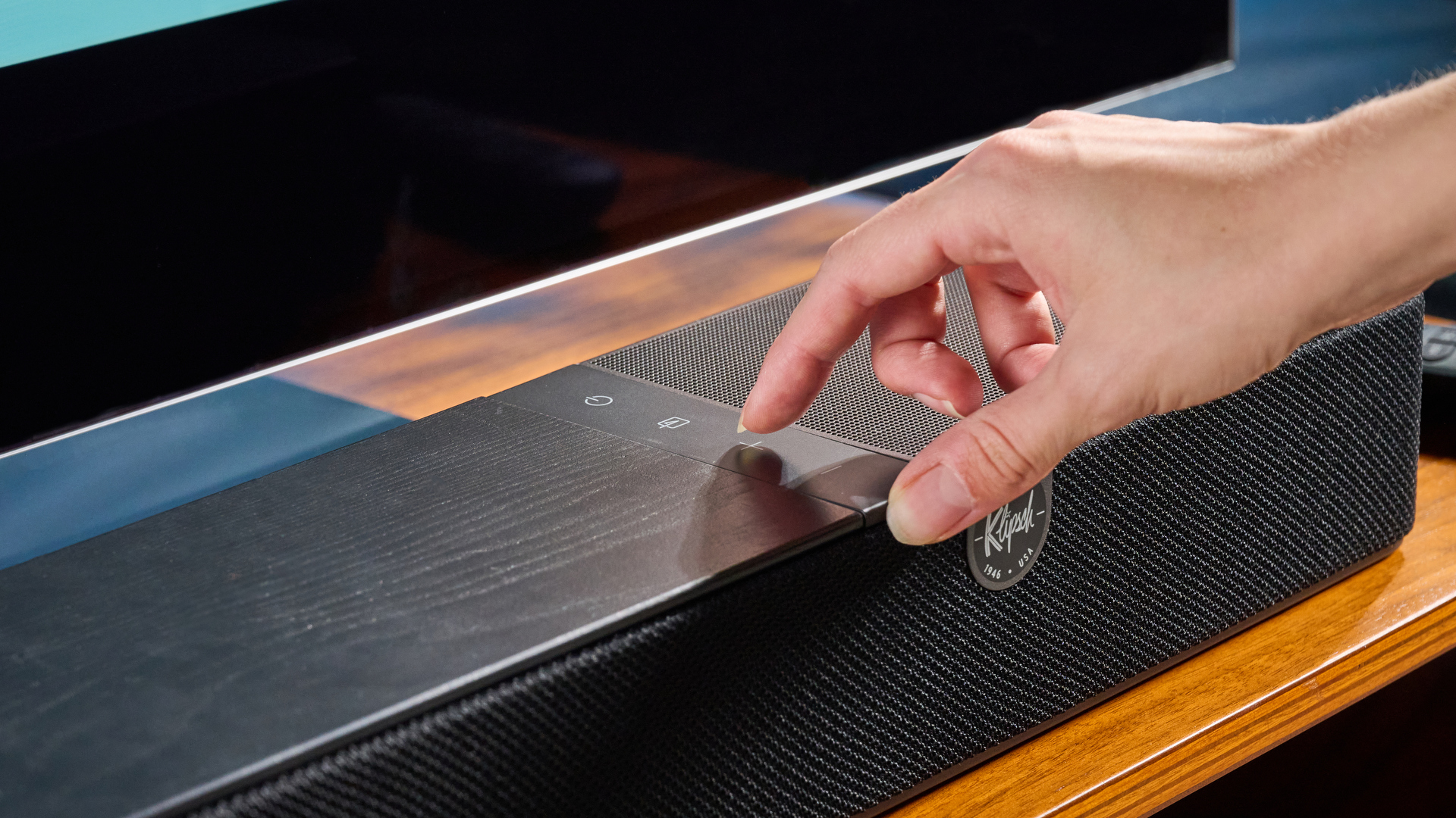 the klipsch flexus core 100 soundbar photographed on a wooden tv stand with a tv behind, showing the remote, buttons, and inputs