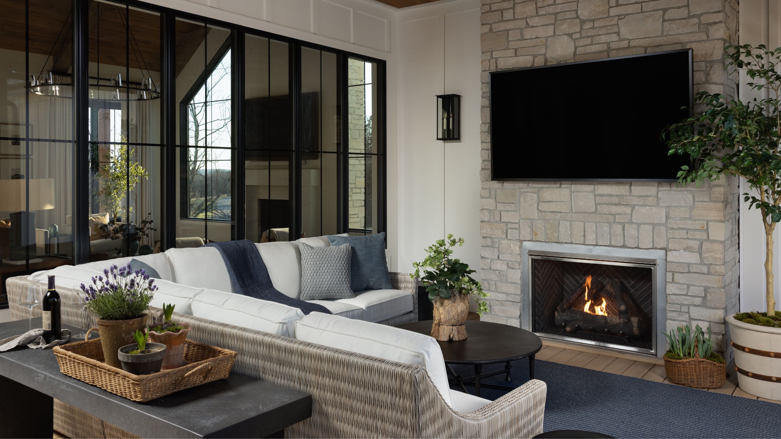 Cozy outdoor living room featuring a woven white sectional, stone stacked fireplace with mounted TV, and large glass windows overlooking nature.