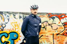 A white man in a navy blue waterproof cycling jacket stands against a graffiti wall