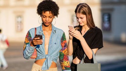 Two young woman are looking at their phone wearing fashionable outfits, this was pictures during fashion month in Italy