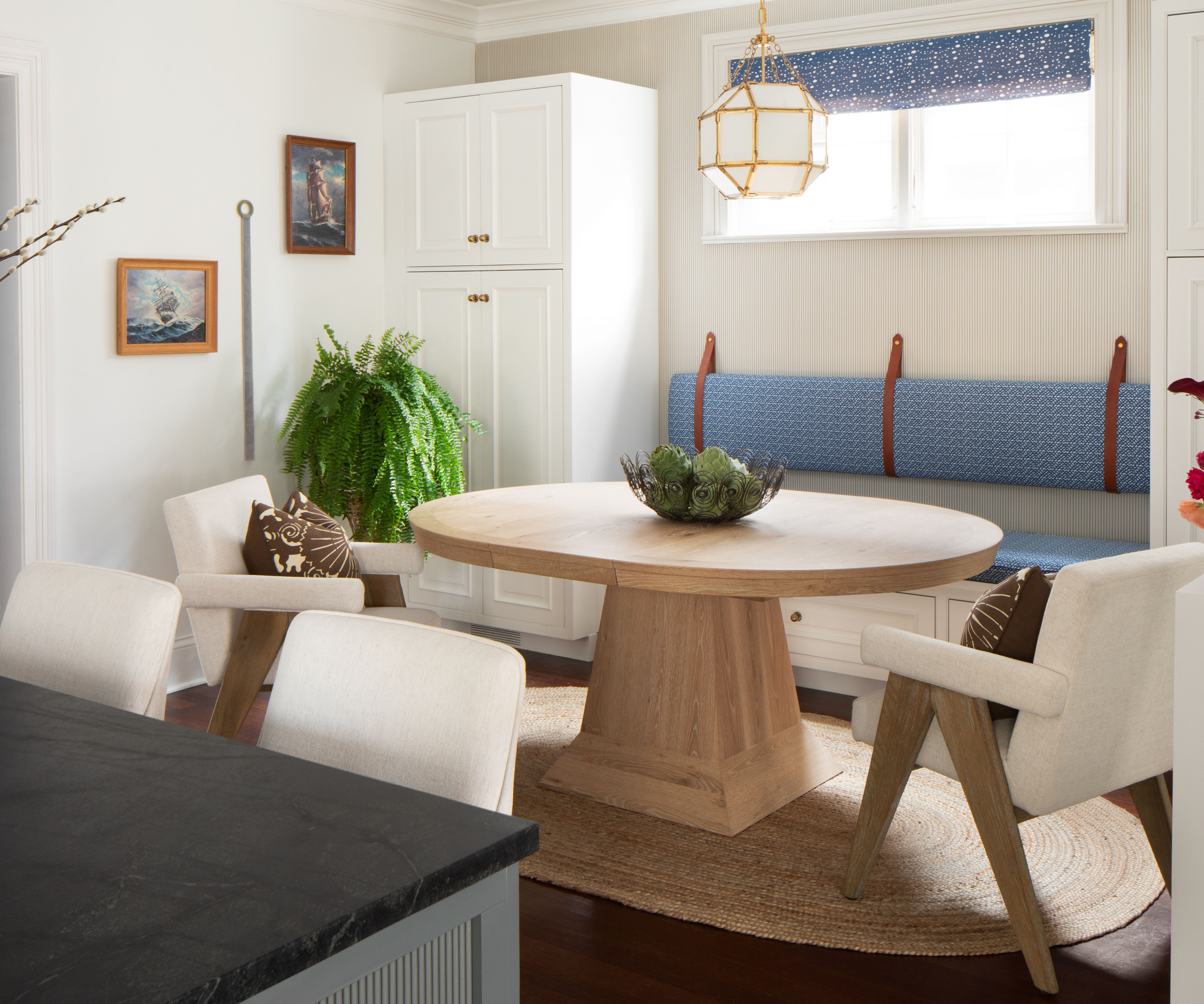 Breakfast nook with a round wood table, low slung armchairs, an upholstered banquette, cream walls, and an antique gold pendant light
