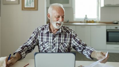 An older man appears happy as he looks over financial paperwork at his kitchen table.