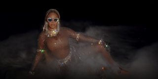 Beyonce sits on the ground while wearing a glittery bodysuit and sequined sunglasses in a scene from 'Black Is King'