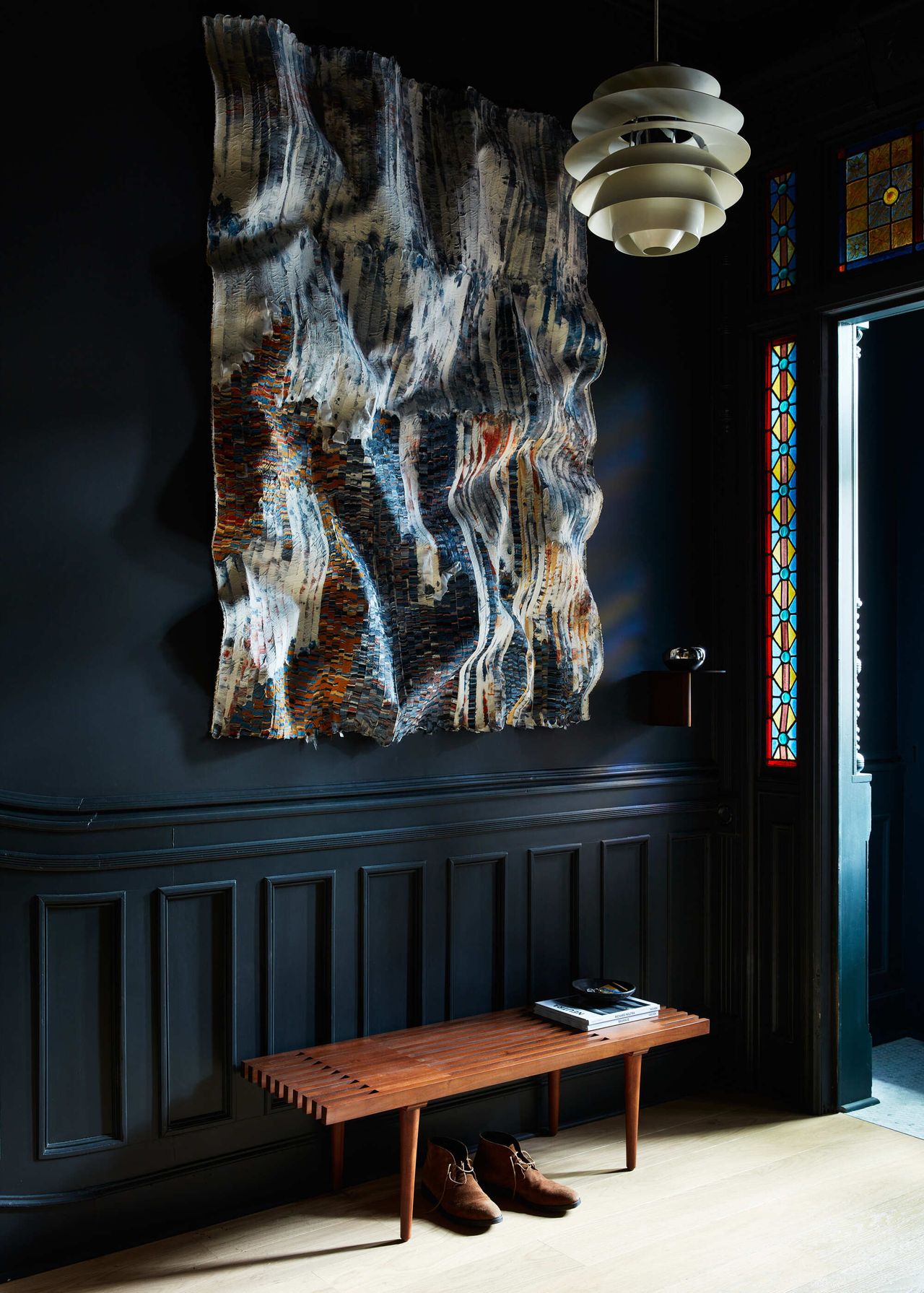 Image of a moody, dark blue entryway with a warm wooden bench. There is a large abstract painting on the wall and a doorway with stained glass.