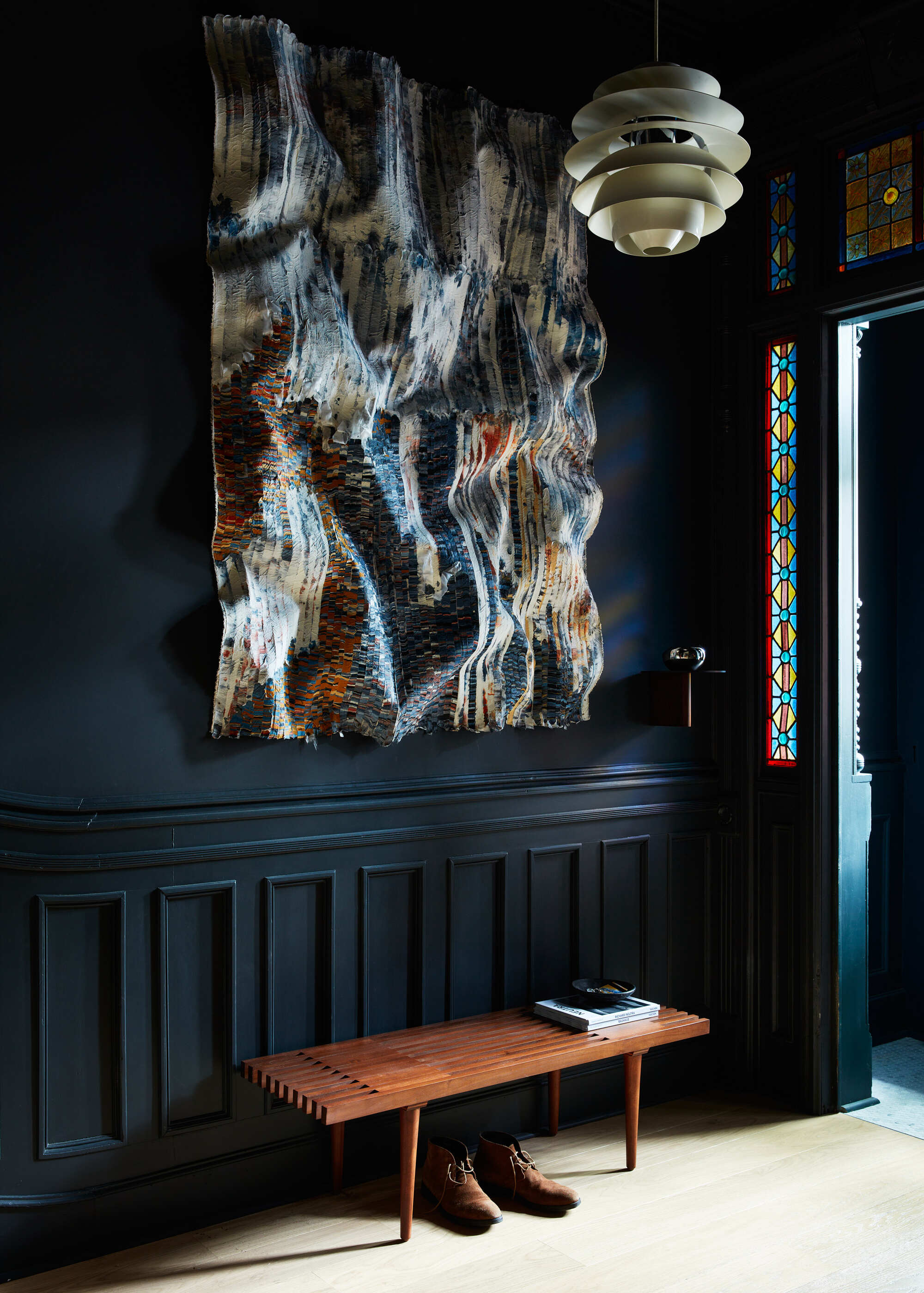 Image of a moody, dark blue entryway with a warm wooden bench. There is a large abstract painting on the wall and a doorway with stained glass.