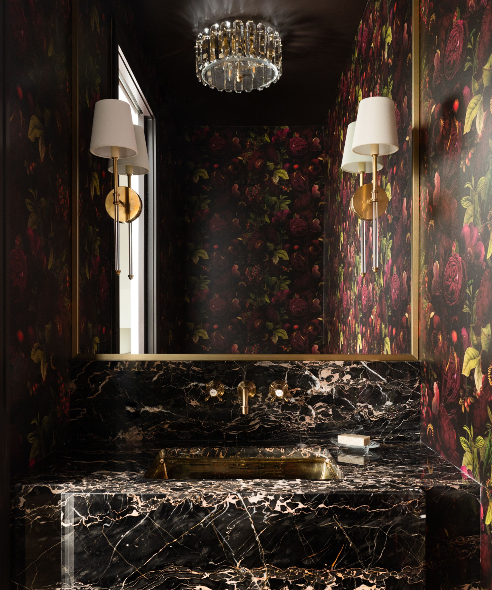 A dramatic powder room with dark floral wallpaper, a black and white veined marble vanity, a gold sink, and elegant brass wall sconces.