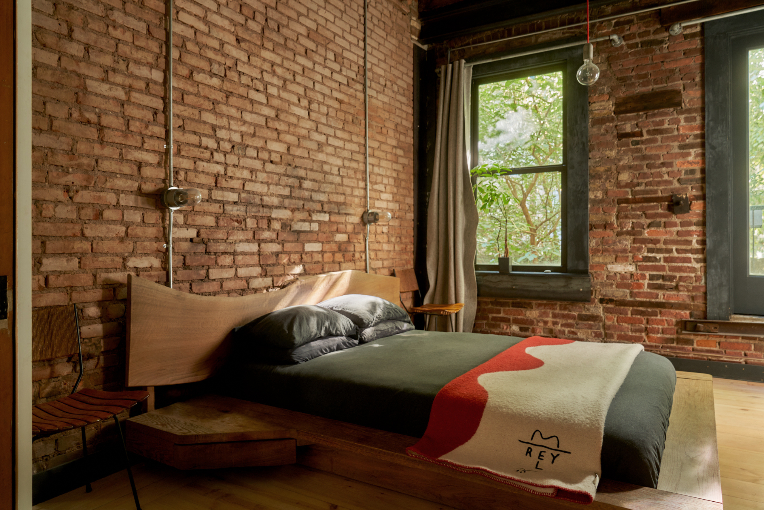 A bedroom with a brick wall, large black framed windows, an extended wooden bed frame, and a gray bedscape with a red and cream throw