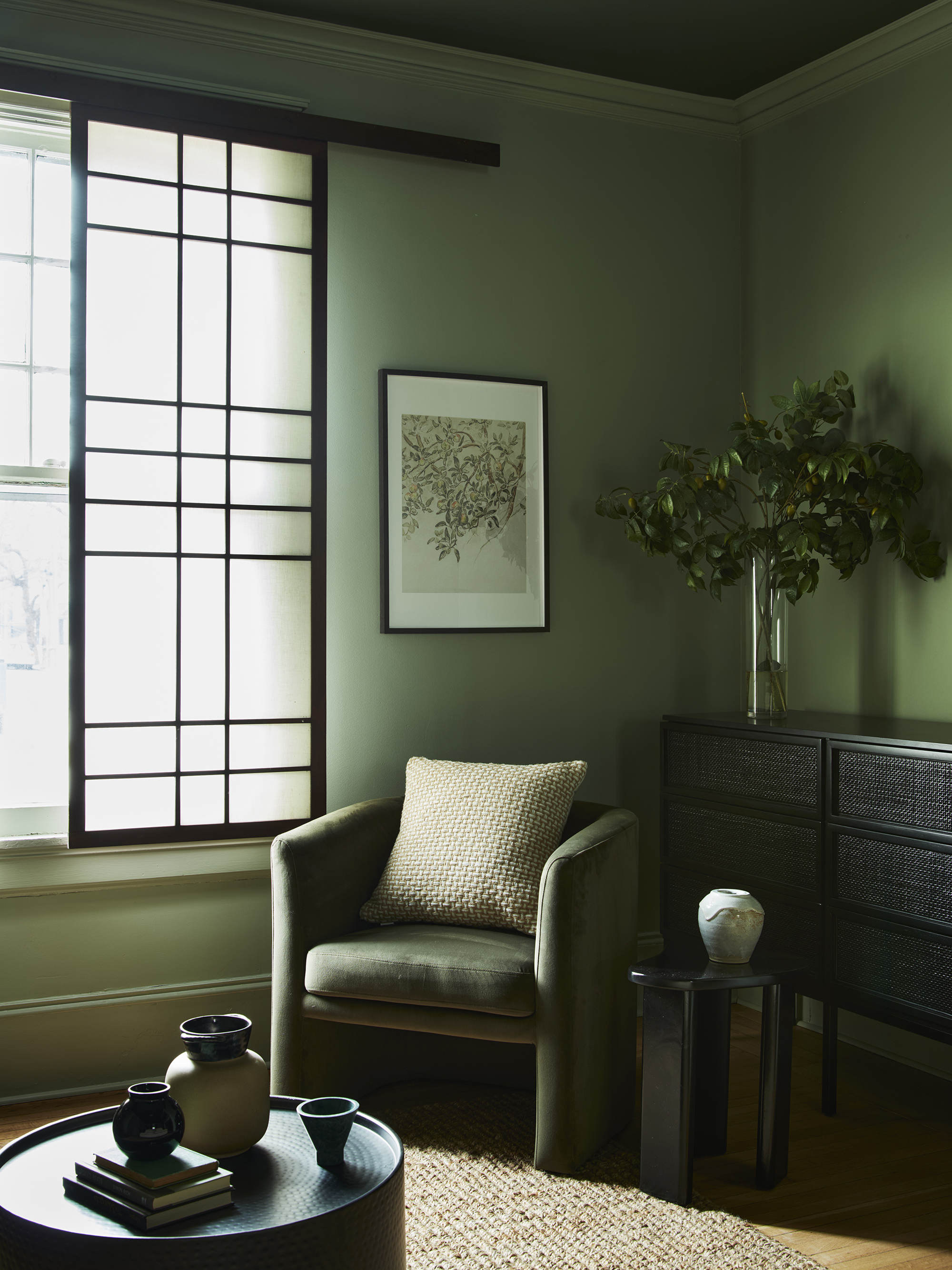 sage green living room with screen over window, green velvet armchair, a console in the corner with a vase with foliage, an artwork hanging on the wall above the chair, a jute rug, and a round black coffee table styled with vases and books