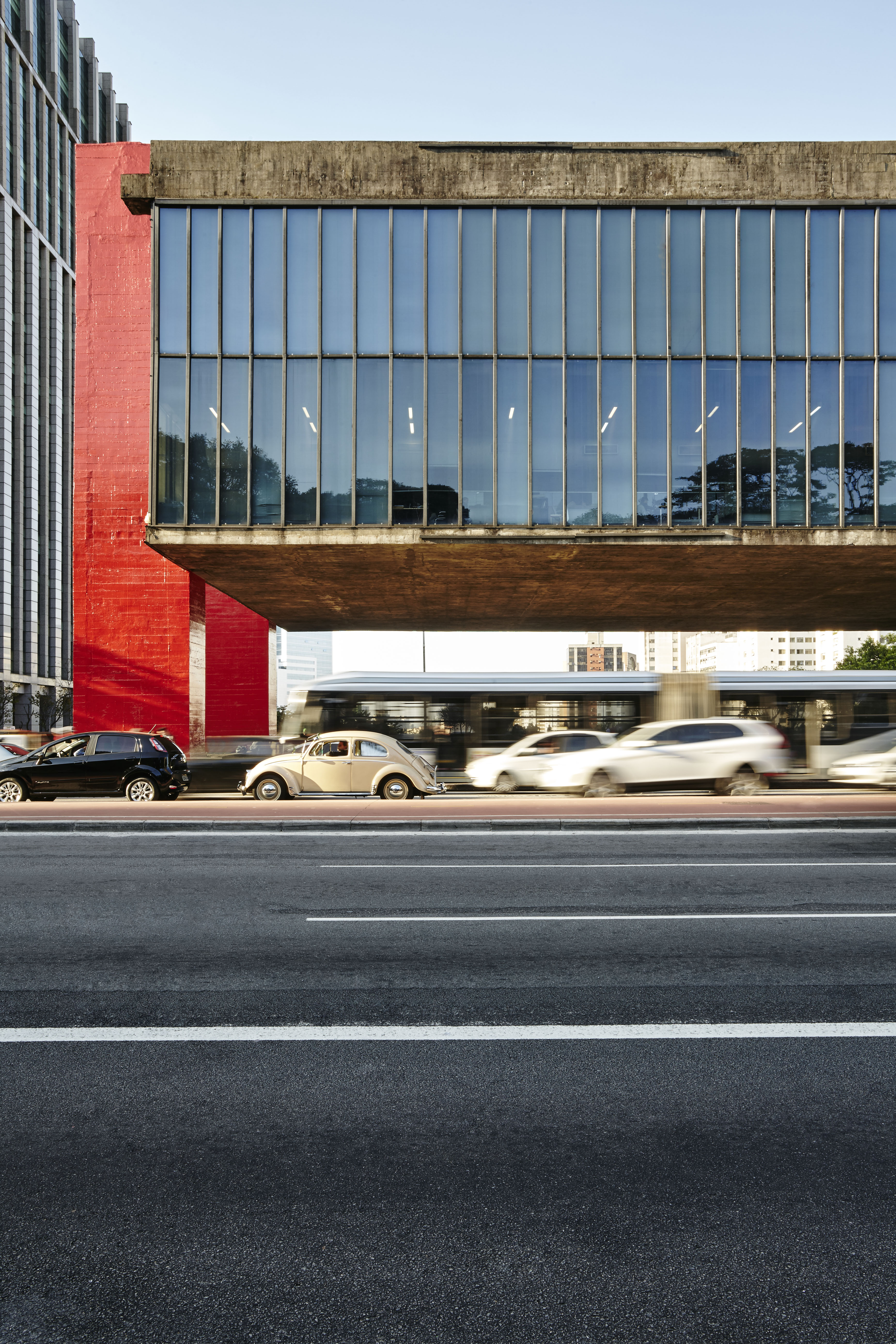 A steel, concrete, and glass museum&#039;s facade positioned on a trafficked road reflects the blue sky on a sunny day as its left hand side&#039;s red-painted columns come to life in the light.