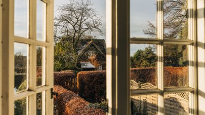 An old style wooden framed window, open, with a view of a cottage and a winter leafless tree. Brown hedges