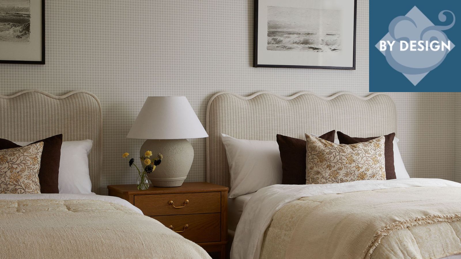 Two beds with scallopped headboard, matching white and cream bedding and brown and floral thorw pillows. Faint checkered wallpaper with framed pictures hung above beds. Bedside table inbetween