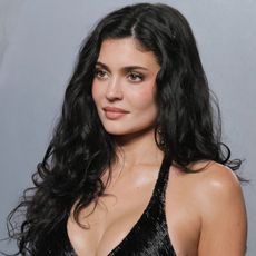 a white woman with long, dark hair poses in front of a gray background