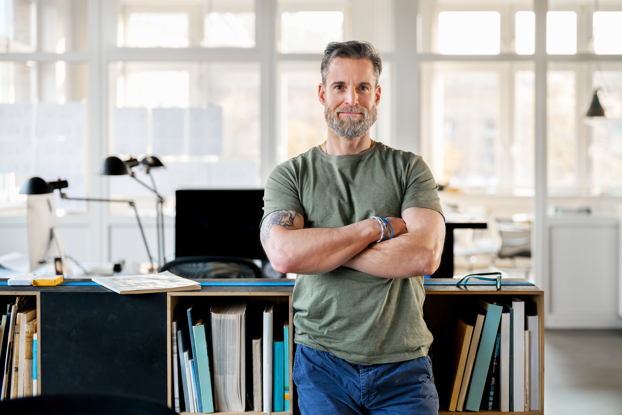 A mature creative director stands with his arms crossed, radiating confidence and experience in a sun-drenched office. The open shelves and large windows in the background emphasise the professional yet approachable atmosphere. Because he works in a creative field, his job is more likely to be impacted by AI.