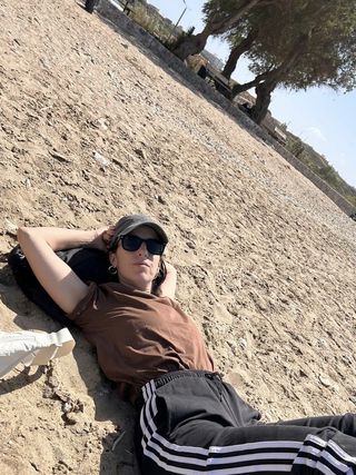 Esta Plakokefalou laying on sand wearing a hat, brown top and striped pants.