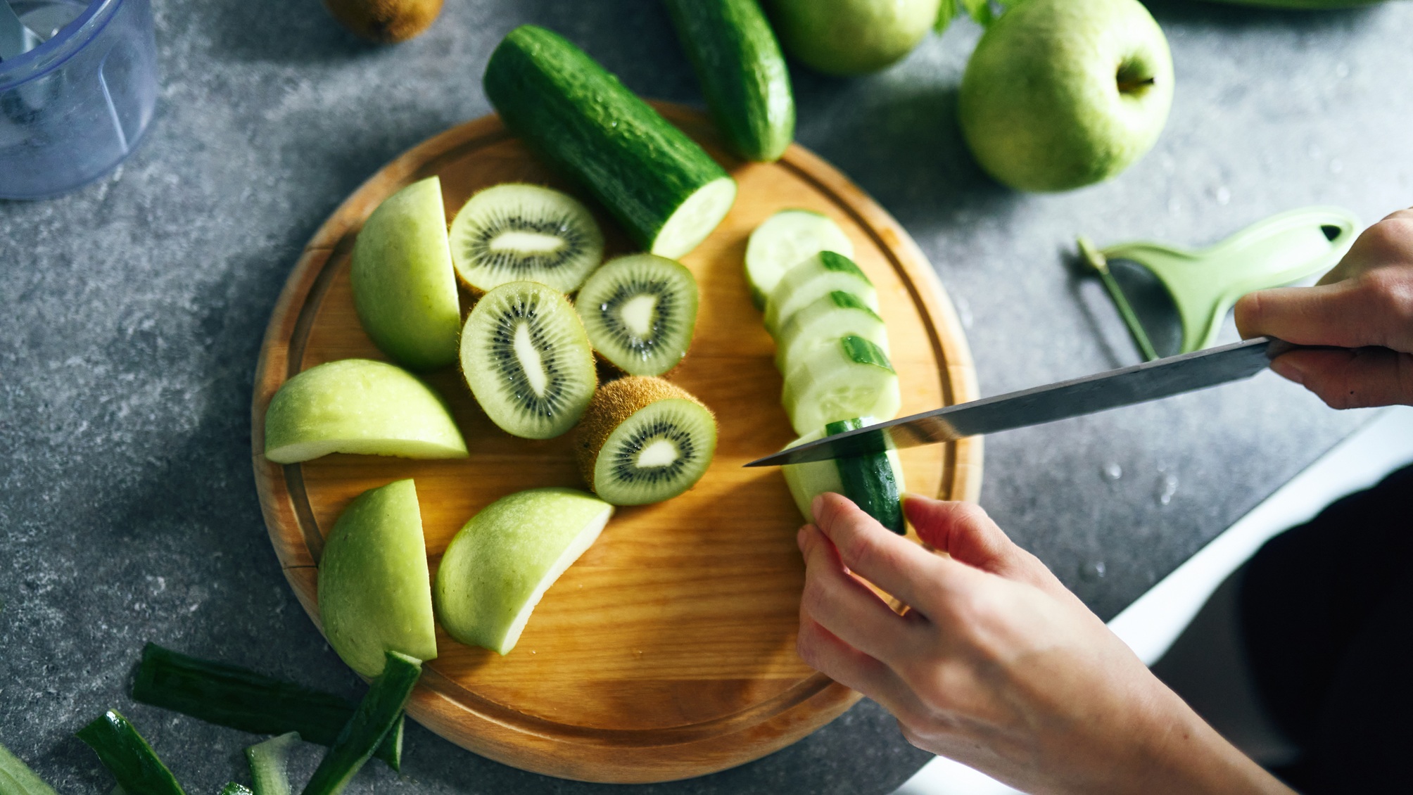 Hands slicing a cucumber on a cutting board with kiwi and green apple.