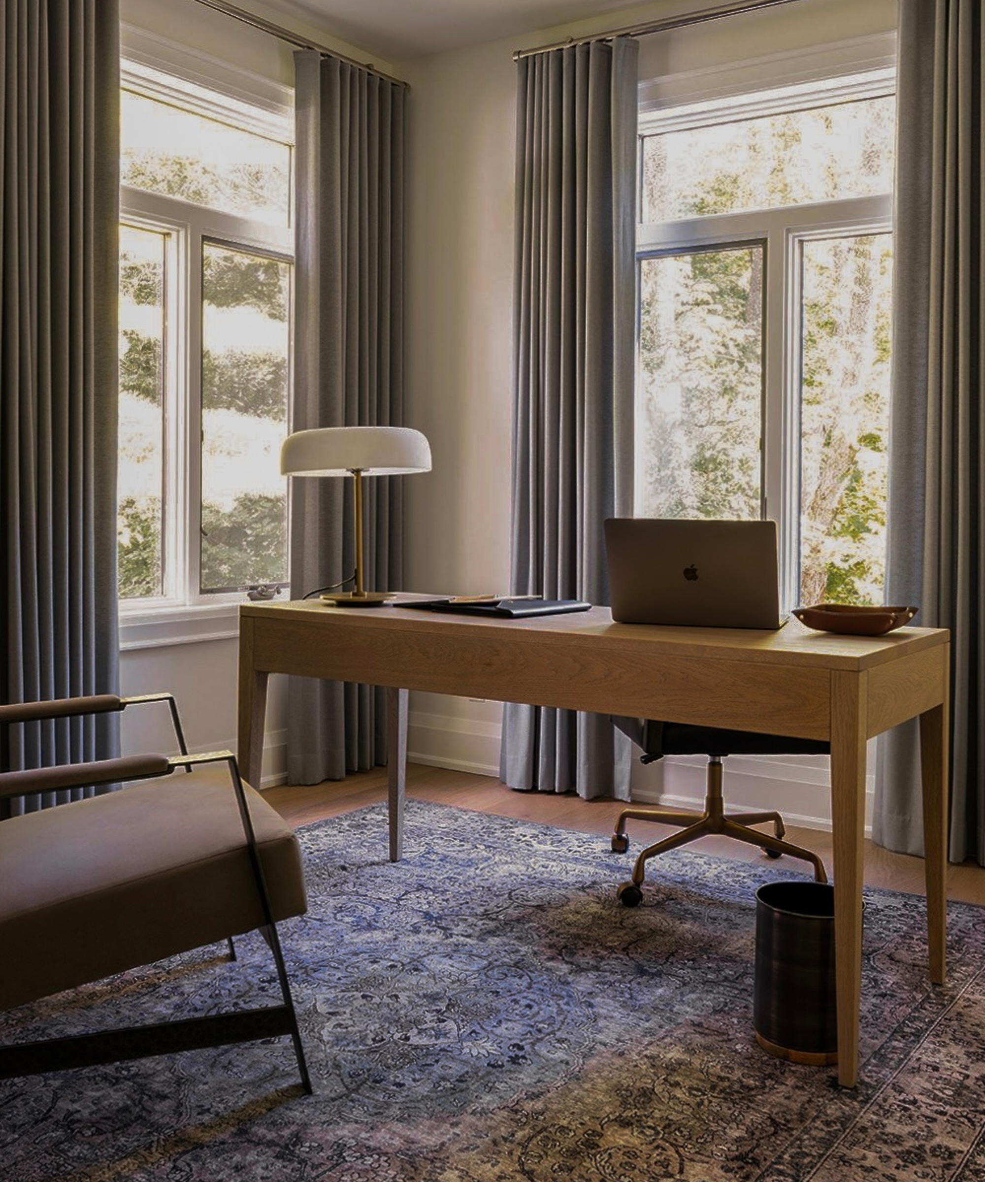 a simple home office with large windows covered by floor to ceiling drapes, a simple wooden desk upon which rests a lamp, a laptop, and papers, an accent chair, a small garbage can, and a persian rug on the floor