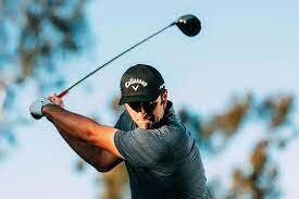 A man wearing a Callaway hat tees off on a golf green