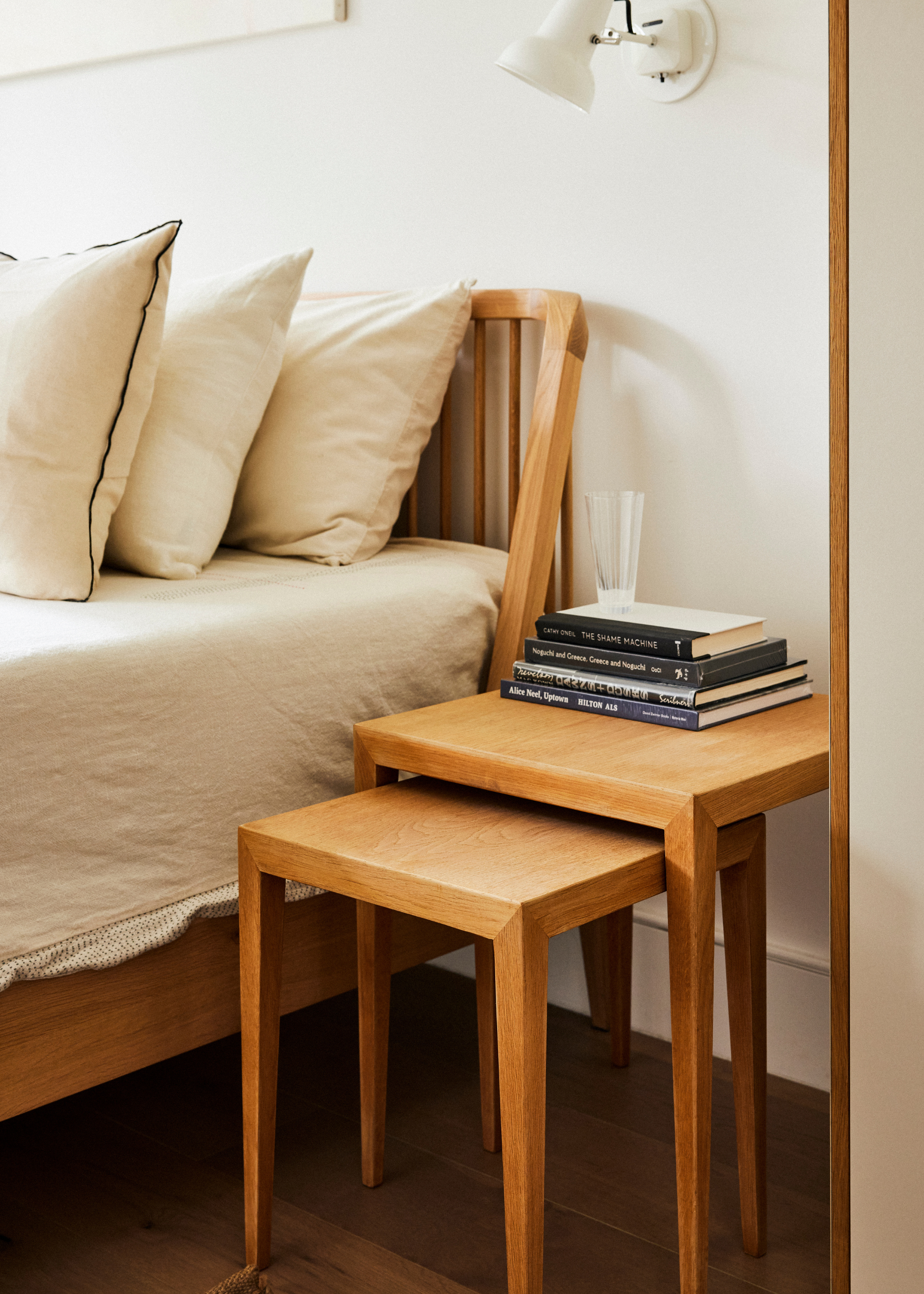 A close-up of a white bedroom corner with a pair of wooden stackable side tables, a stack of books, and a glass
