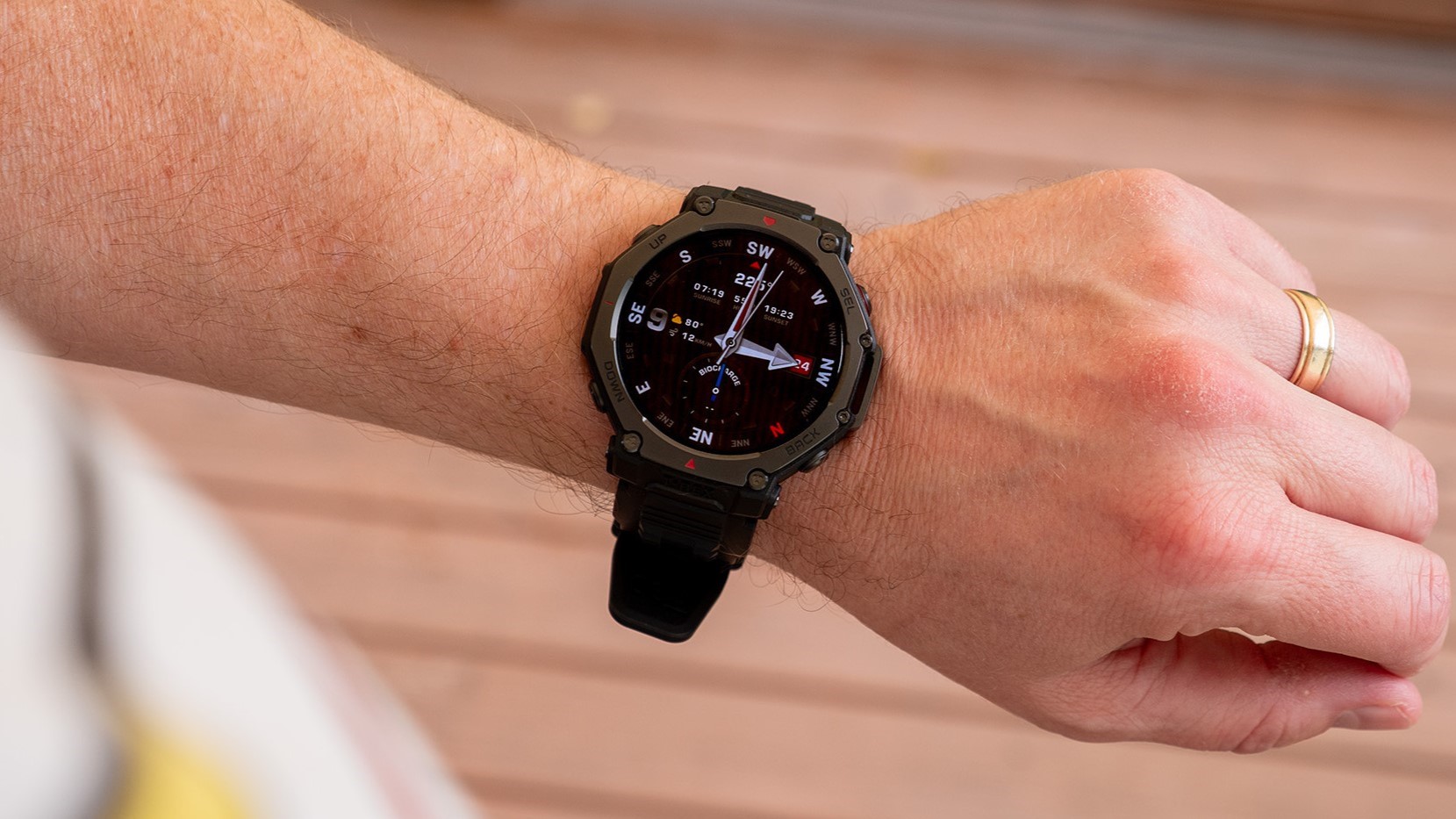 A photo of the Amazfit T-Rex 3 Pro on a man's wrist above a wooden deck, showing a standard watch face.