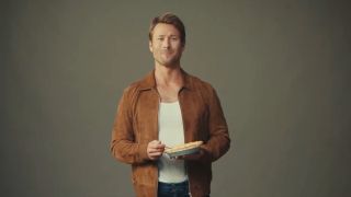 Glen Powell smiles as he holds a fork and a pie in a Hit Man promo video.
