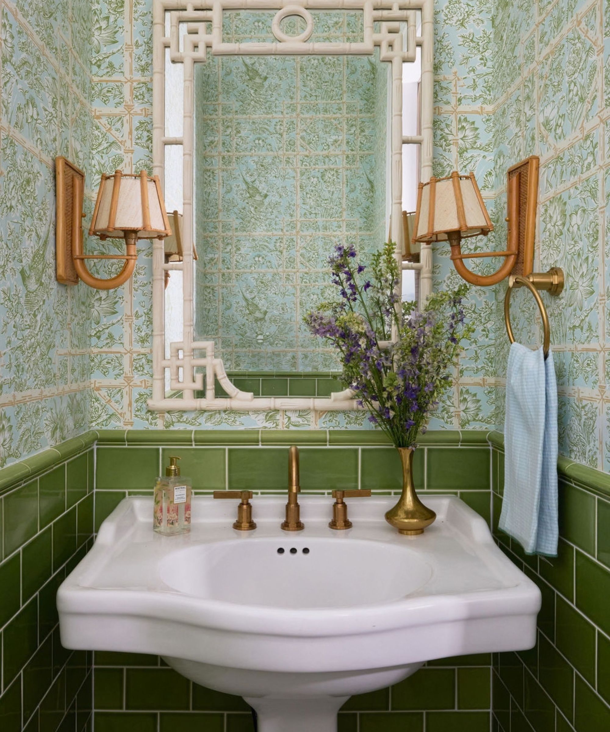 Powder room with green subway tile, light green floral wallpaper, a white pedestal sink with brass hardware, and a bamboo mirror