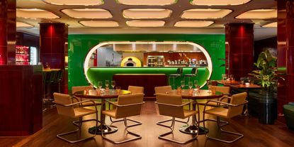 An immersive restaurant with coffered, backlit ceilings, an oblong, lacquered, green kitchen, and leather and chrome chairs boasts cherry plum columns in curved ceramic tiles and a warm atmosphere.