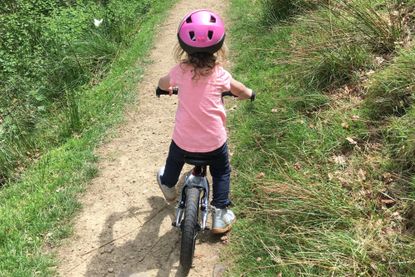 Best balance bikes image shows a child in a pink helmet and t-shirt, black leggings and silver boots riding a balance bike with their back to the camera