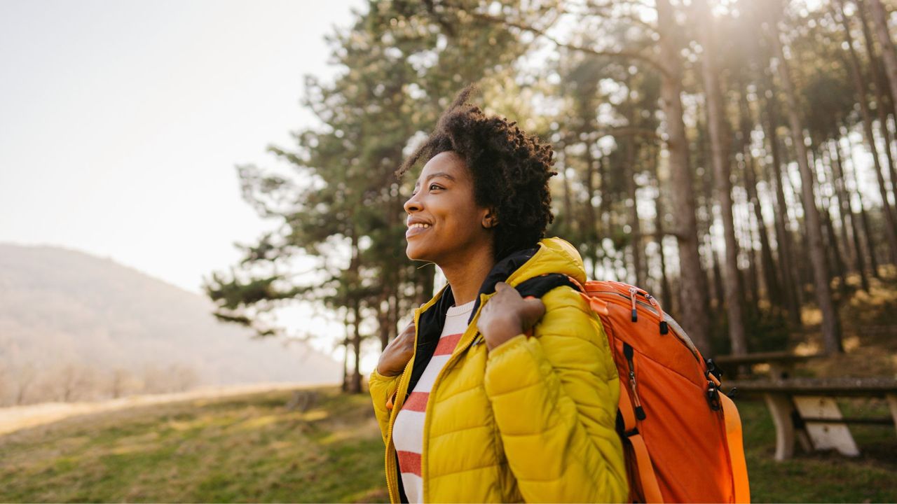 Woman taking a break from walking intervals in an autumnal forest, wearing jacket and bright orange backpack