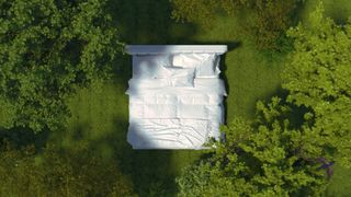 A white bed frame with a mattress and white bedding made on it in the middle of a green field surrounded by trees