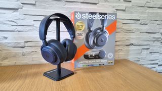 The SteelSeries Arctis Nova Pro Omni headset sitting on a headset stand in front of its box and a white brick background and on a wooden surface.