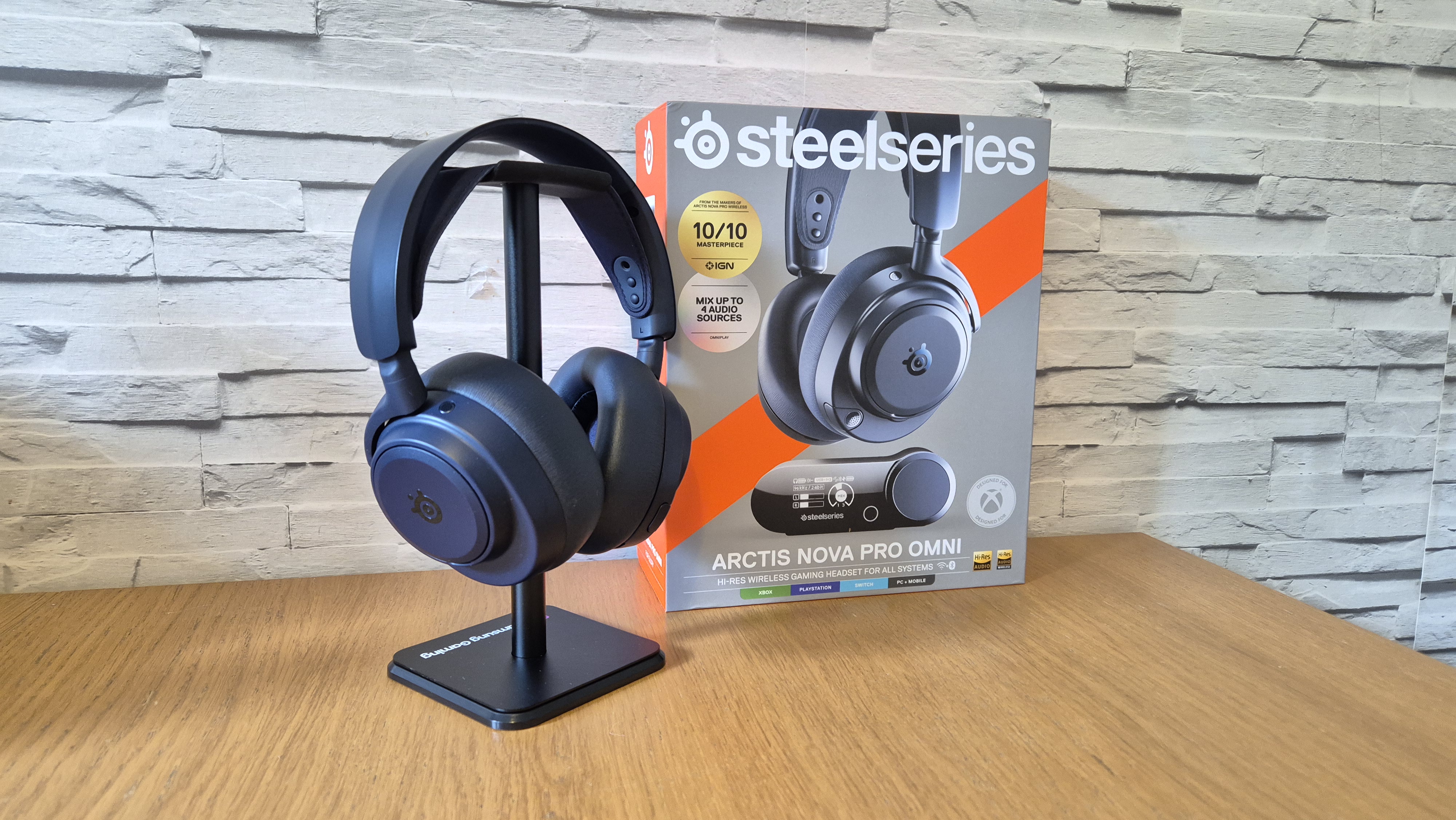 The SteelSeries Arctis Nova Pro Omni headset sitting on a headset stand in front of its box and a white brick background and on a wooden surface.