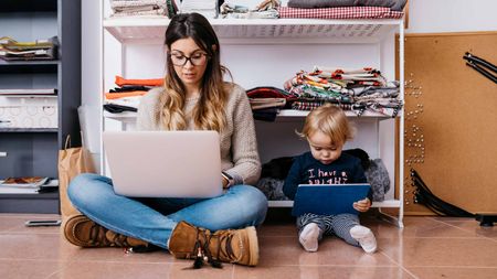 A mom works on her laptop with her toddler looking at a tablet next to her.