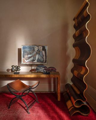 A sun-lit room features a red carpeted floor, a minimalist wooden desk covered in artworks, a wrought iron and leather mid-century stool, and a wavy room divider casting shadows on the walls.