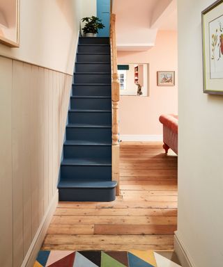 Pale pink painted hallway with tongue and groove paneling, dark blue painted stairs, light wood flooring and multi colored geometric tiled flooring, framed painting on wall