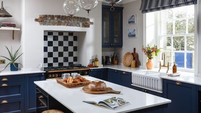 A blue and white kitchen with blue cabinets, a white kitchen island, and a black and white tiled backsplash behind a black stove. On the kitchen island is a loaf of bread, a tray with breakfast goods, and an open recipe book. To the right is a white fluted sink with gold faucets next to a vase of flowers, below a bright and sunny window.
