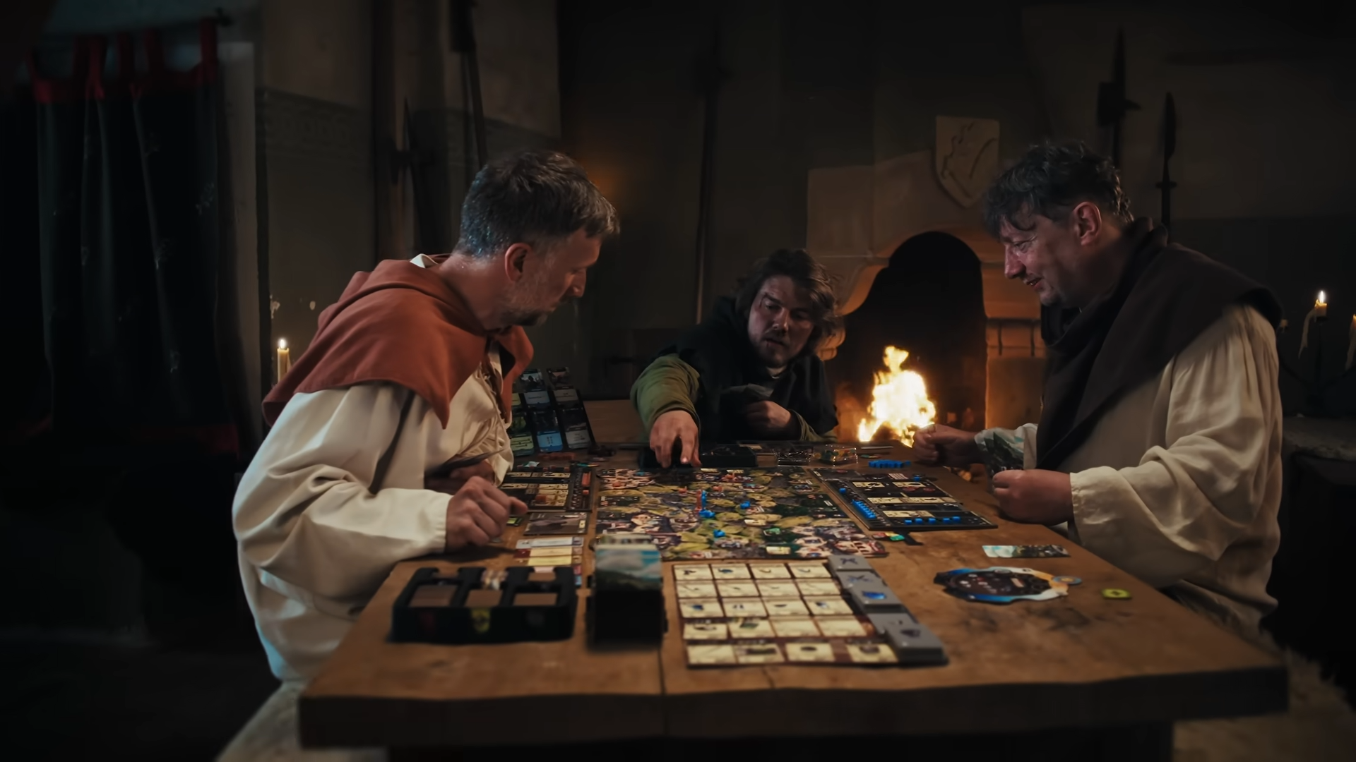 Three men in medieval outfits sit in a castle-like environment, playing the Kingdom Come: Deliverance board game