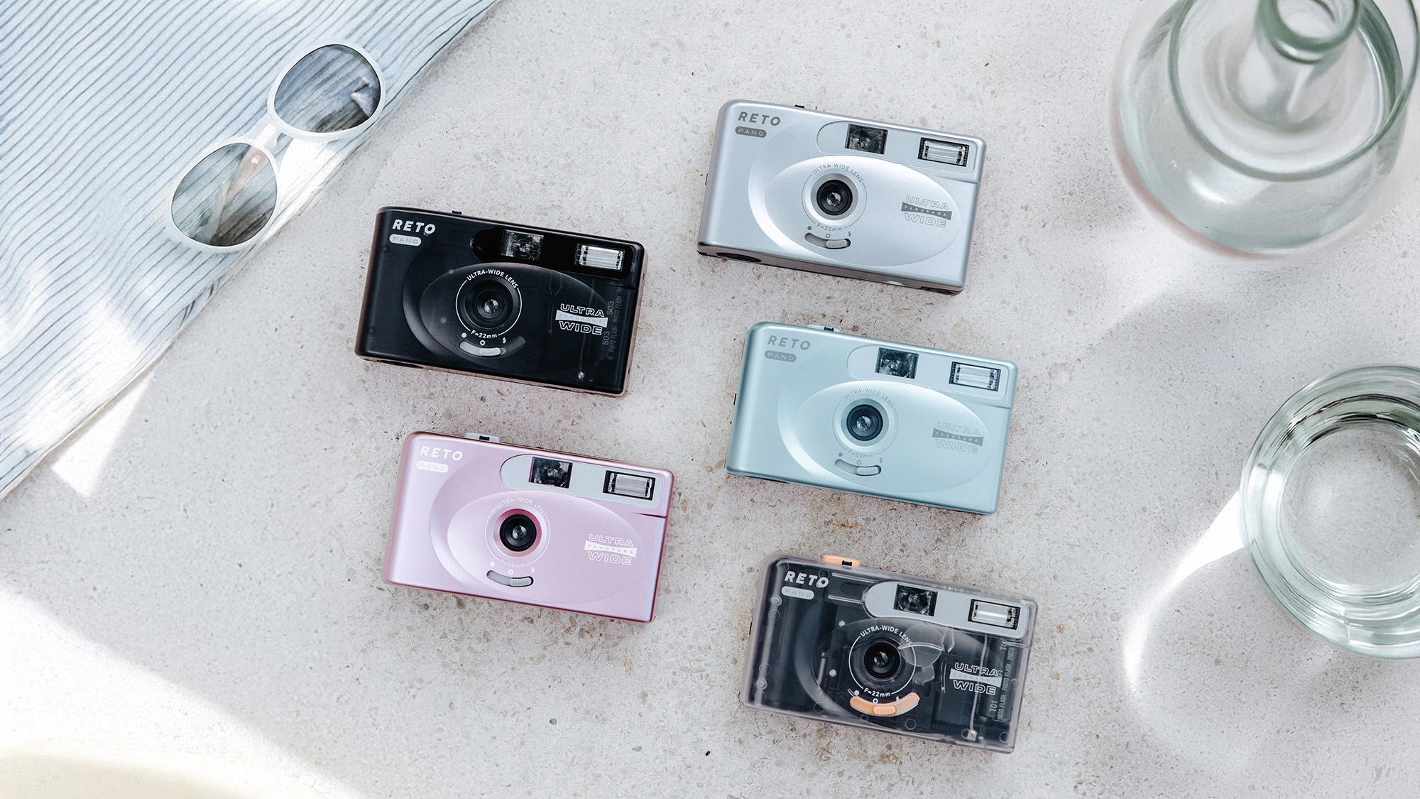 Top-down Reto Pano lifestyle image of all five Reto Pano cameras on a marble surface next to glass vessels and sunglasses