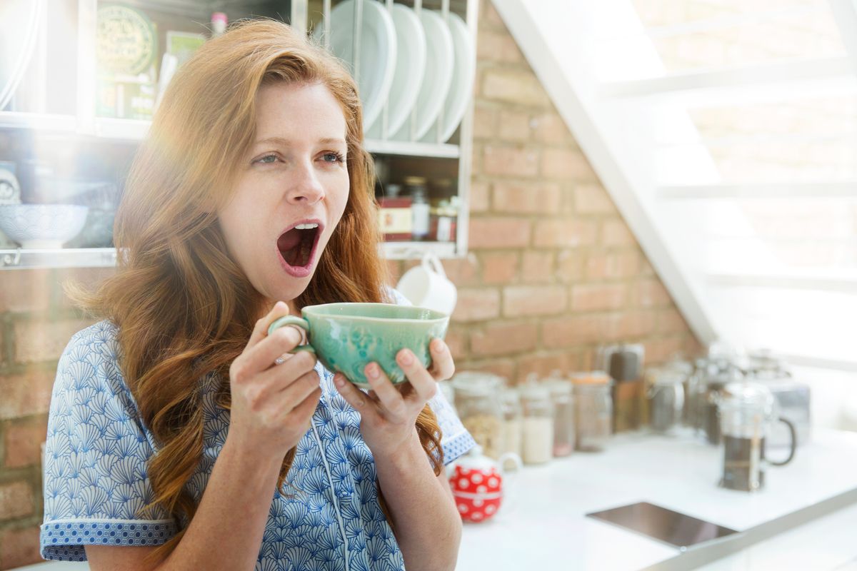 A woman with red hair yawns while holding a large mug with both hands