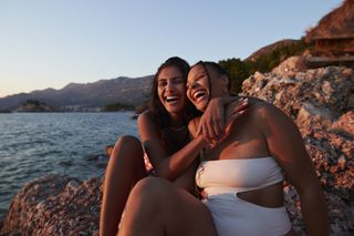 Two friends giggling while wearing swimwear on a beach in the evening sun.