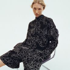 Blonde woman wearing floral white and black playsuit, sat on chair with white background.